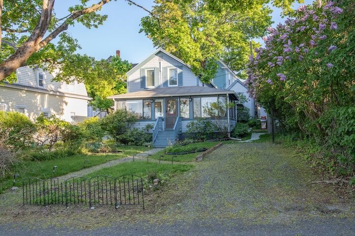 White house with porch and front yard, framed by green trees and purple blossoms, beside a gravel driveway