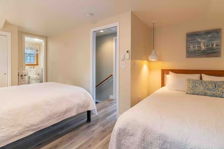 Bright bedroom with two white beds, wood floors, and a small adjoining bathroom.