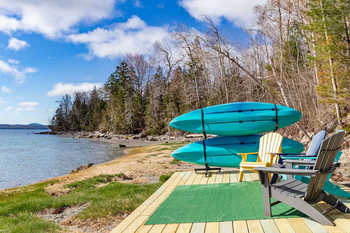 Lakeside dock with turquoise kayaks, Adirondack chairs, and a green mat under a blue sky