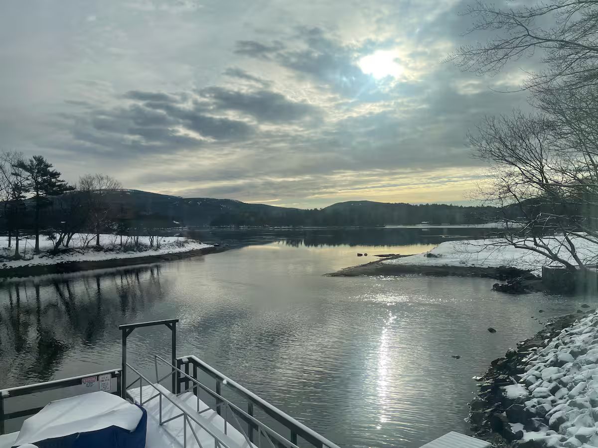 Snowy river landscape at dusk with calm water reflecting a cloudy sky and a small dock in the foreground