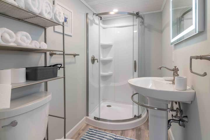 Small white bathroom with shower stall, toilet, sink, mirror, and towel shelves