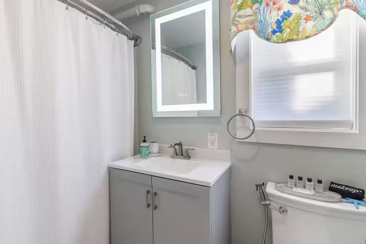 Bright bathroom with gray vanity, mirror, shower curtain, and white toilet beside a small window.