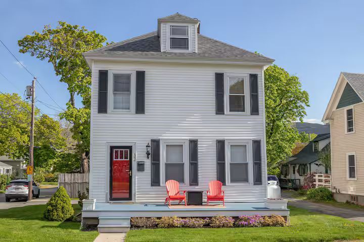 White two-story house with a red front door, black shutters, and a small porch with two red chairs.