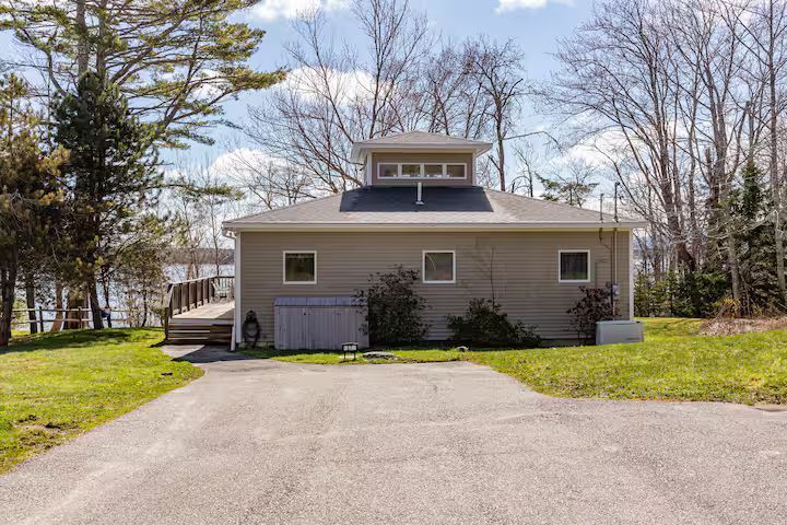 Small gray house with a driveway, front lawn, and trees under a partly cloudy sky