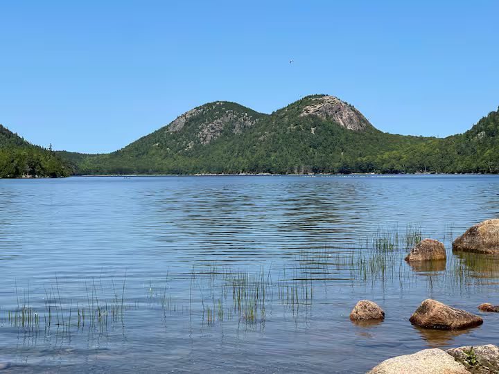 Calm lake with rippling water, green hills in the distance, and rocks along the shore under a clear blue sky
