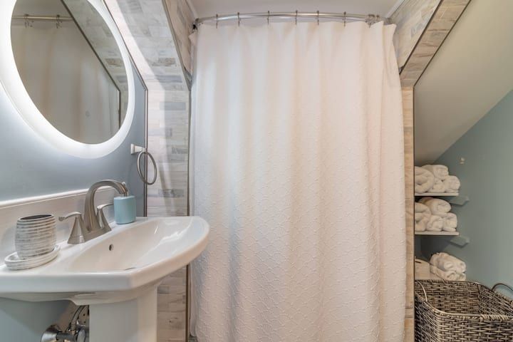Small bathroom with white sink, round mirror, shower curtain, and shelves of folded towels