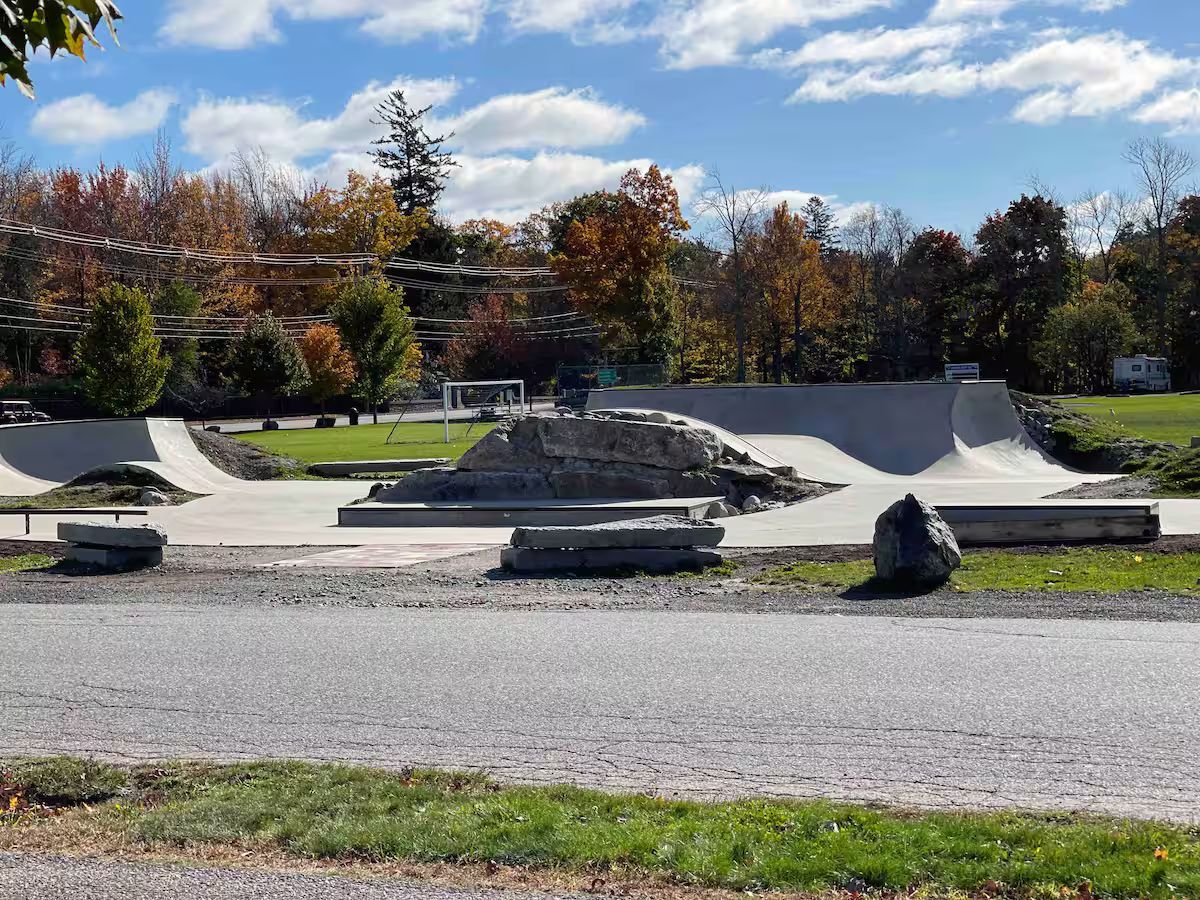 Outdoor skatepark with ramps and rails under a blue sky, surrounded by autumn trees.