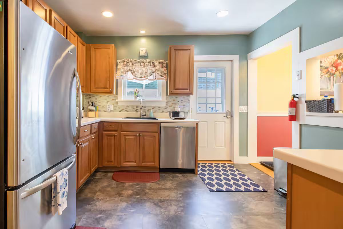 Bright kitchen with stainless steel fridge, wood cabinets, tiled floor, and a back door with a patterned mat.