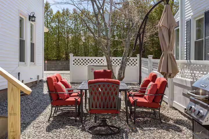 Patio seating area with red cushioned chairs, black table, gravel ground, and a closed umbrella beside a grill.