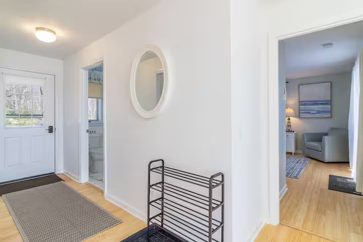 Bright entryway with white walls, wood floors, mirror, and hallway leading to a bedroom and bathroom.