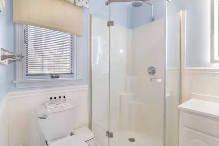 Bright white bathroom with a corner glass shower, toilet, window, and vanity sink.