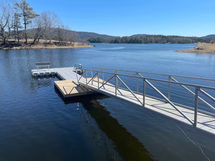 Dock extending into a calm blue lake with a metal ramp, under a clear sky and distant tree-lined shore