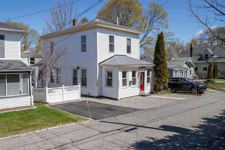 White two-story house with a front porch, driveway, and parked car on a sunny residential street
