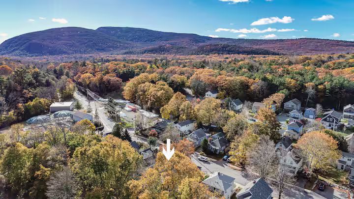 Aerial view of a small town with autumn trees and mountains in the background, with a white arrow pointing down.