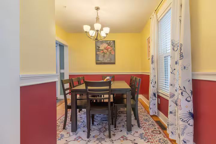 Dining room with yellow walls, red wainscoting, dark table and chairs, chandelier, and floral curtains.