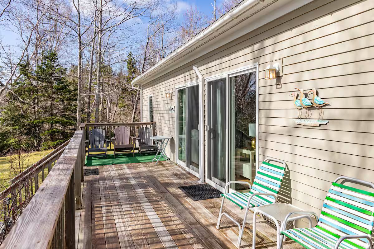 Wooden deck beside a beige house with sliding doors, green chairs, and a bench overlooking trees