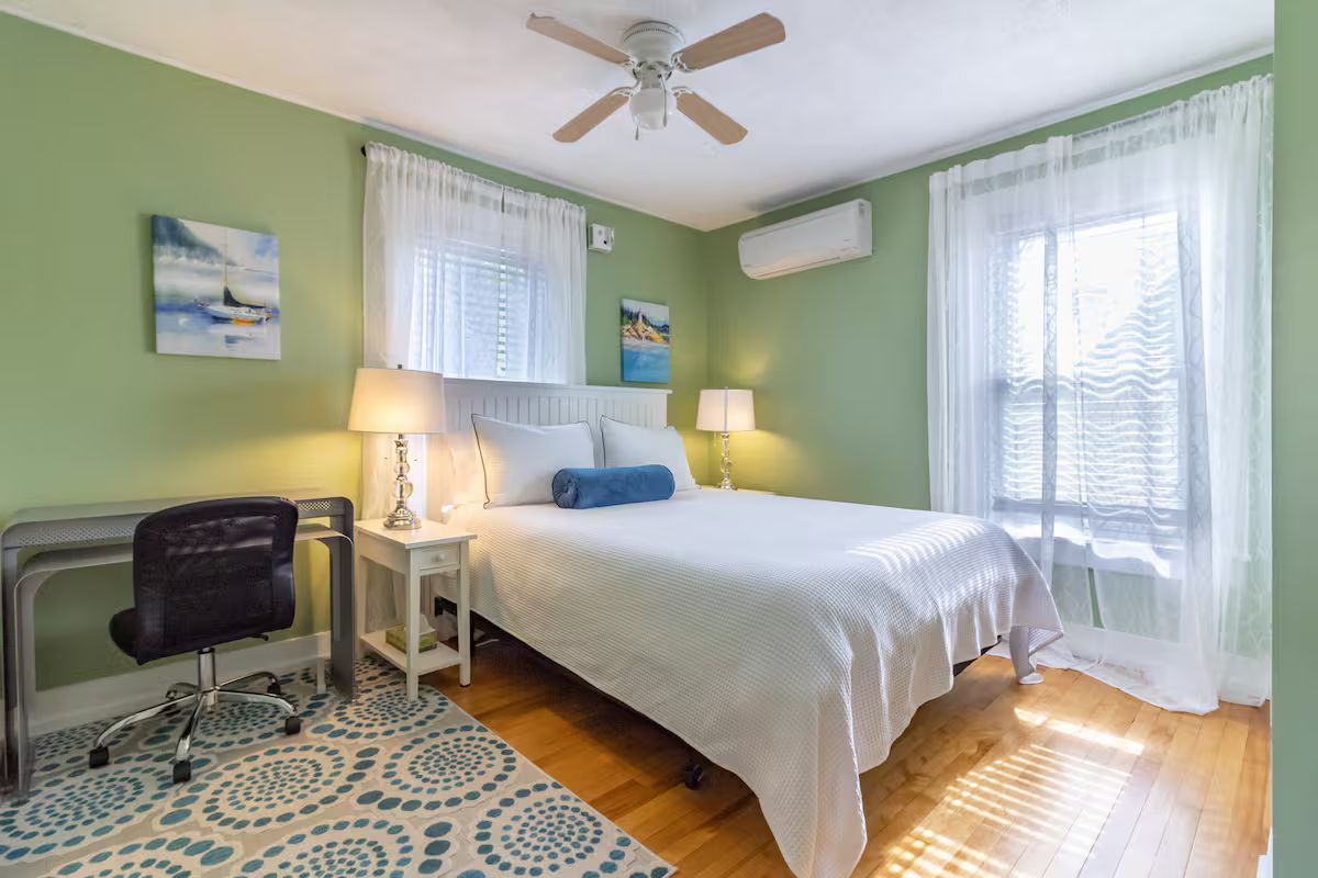 Cozy green bedroom with white bed, desk, patterned rug, and sunlight through sheer curtains