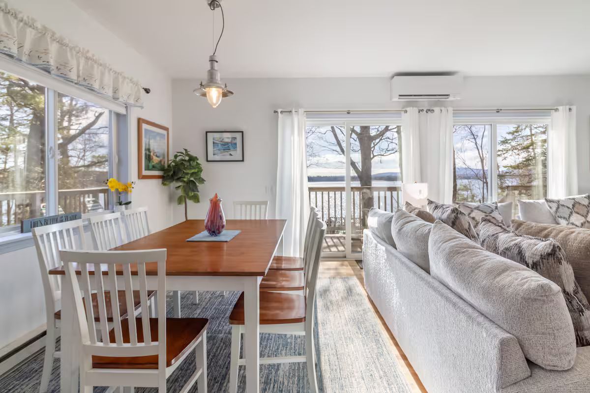 Bright dining and living room with wooden table, white chairs, gray sofa, and large windows