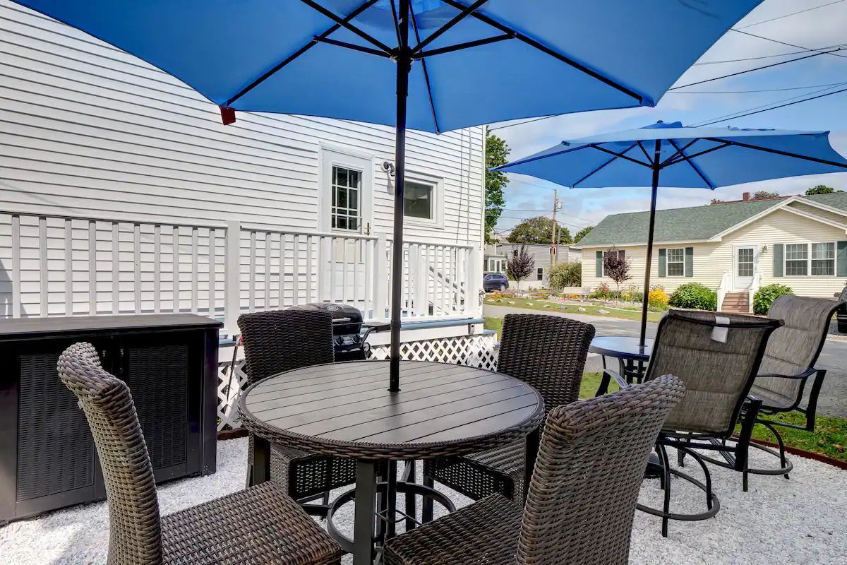 Patio seating area with round table, black wicker chairs, and blue umbrellas beside a white house