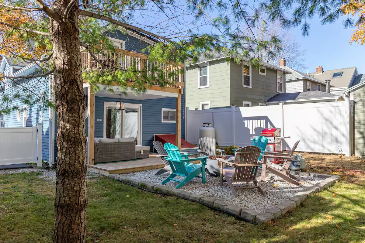 Backyard patio with colorful chairs, fire pit, white fence, and two-story house in the background