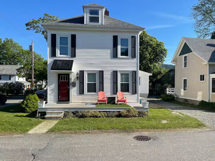 Two-story white house with black shutters, red door, and two red chairs on the front porch