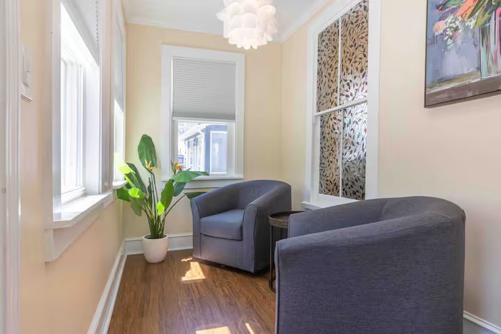 Narrow sunlit hallway seating area with two dark gray chairs, a potted plant, and wood floors