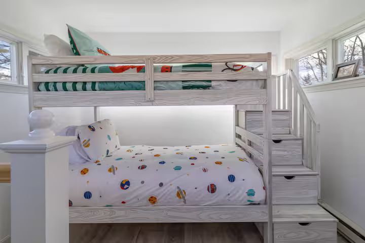 White bunk bed in a bright bedroom with a lower twin bed and staircase drawers on the right