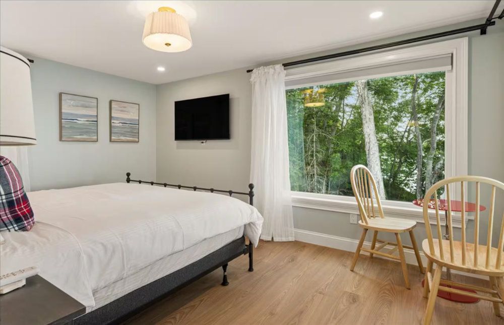 Bright bedroom with queen bed, wall-mounted TV, and a small table by a large window overlooking trees