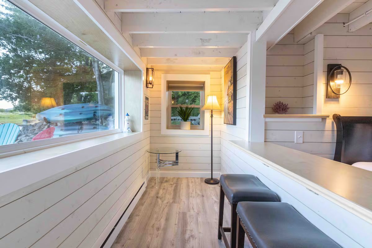 Bright, narrow porch with white paneled walls, wood floor, large window, and dark bench seating.