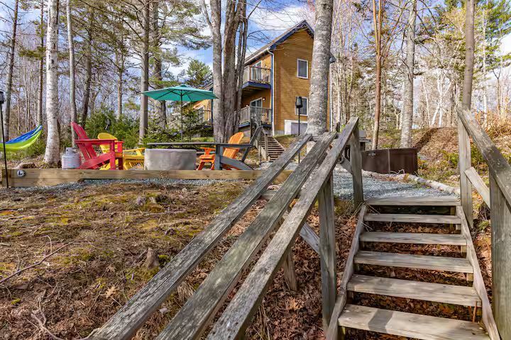 Wooden cabin with deck and stairs in a wooded yard, with patio furniture and a red chair