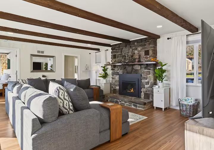 Bright living room with gray sofa, exposed wood beams, stone fireplace, and hardwood floors