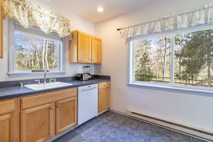 Bright kitchen with wood cabinets, white appliances, blue countertop, and two windows with lace valances