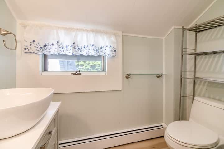 Small bathroom with sink, toilet, towel bars, window, and wire shelving in light gray tones