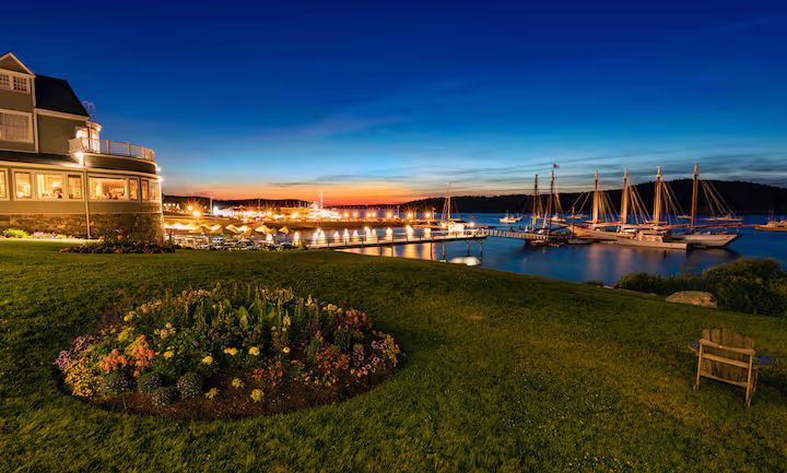 Dusk view of a marina with lit harbor, sailboats, and a grassy flowerbed beside a building and bench