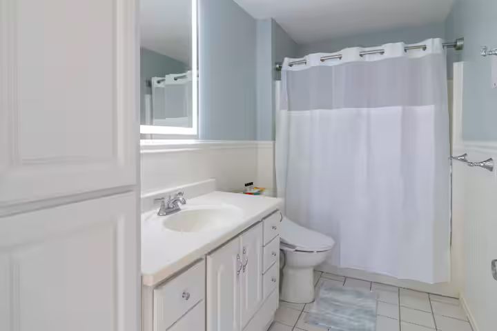 Bright bathroom with white vanity, toilet, mirror, and shower curtain on tiled floor.