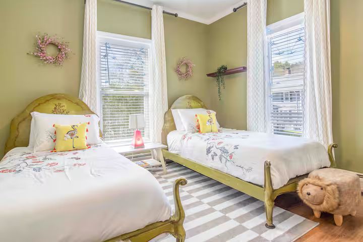 Two twin beds with white bedding in a pale green bedroom, with striped rug and stuffed bear on the floor
