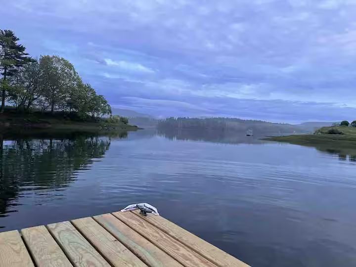 Wooden dock overlooking a calm lake at dusk, with trees and cloudy blue sky reflected in the water.