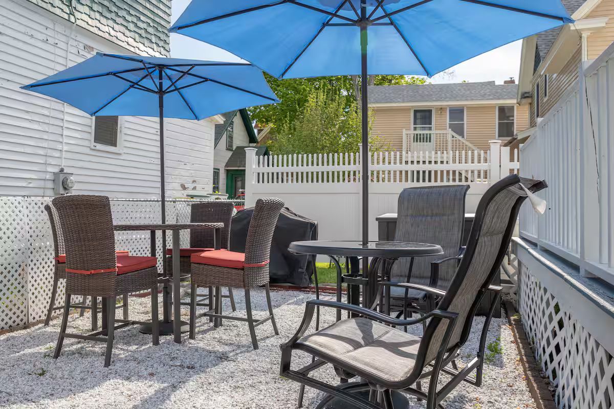 Patio seating with blue umbrellas, wicker chairs, and a small table beside a house and white fence