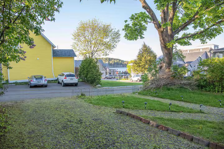 Grassy residential lot with large tree, parked cars, and yellow house on a sunny day