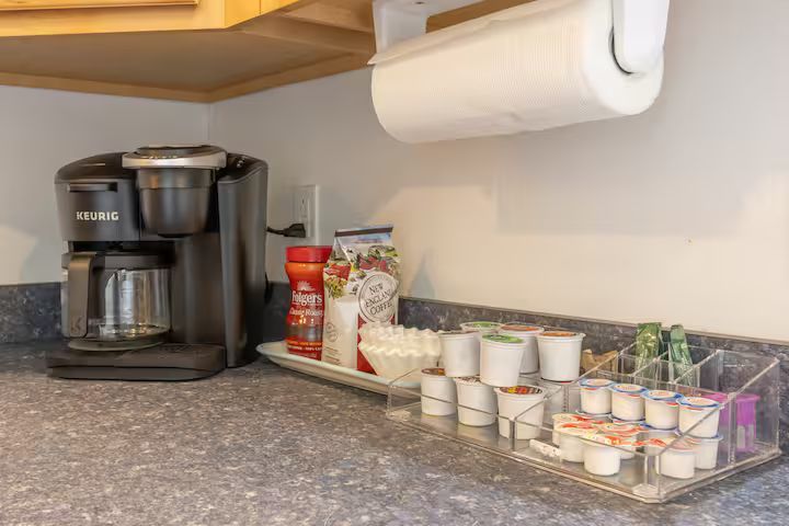 Kitchen counter with coffee maker, pods, mugs, tea boxes, and a paper towel roll underneath a cabinet