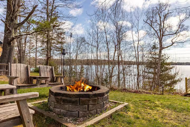 Lakeside fire pit with flames, benches, and trees on a grassy shore under a cloudy sky