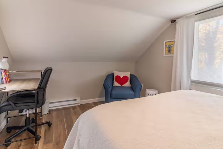 Cozy attic bedroom with a blue chair, heart pillow, desk, and large window.