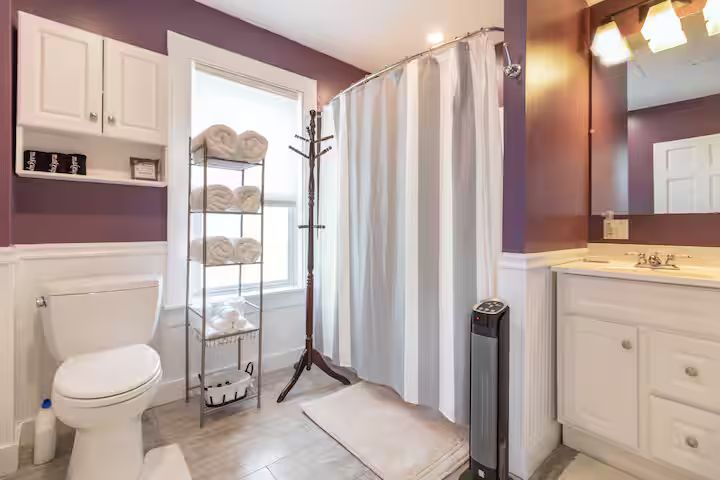 Small purple bathroom with toilet, shower curtain, vanity, and storage shelf by a window.