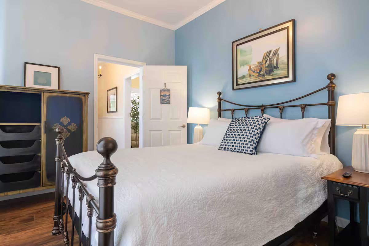 Blue bedroom with a metal bed, white bedding, dresser, lamp, and open door to the hall