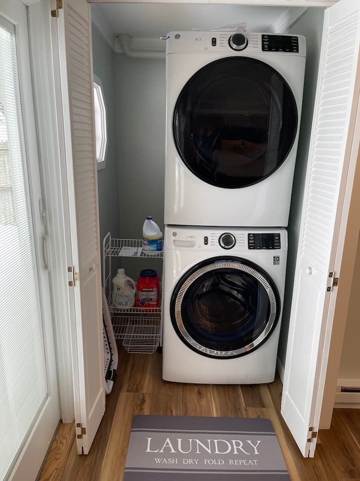 Stacked white washer and dryer in a narrow laundry closet with a “LAUNDRY” mat.