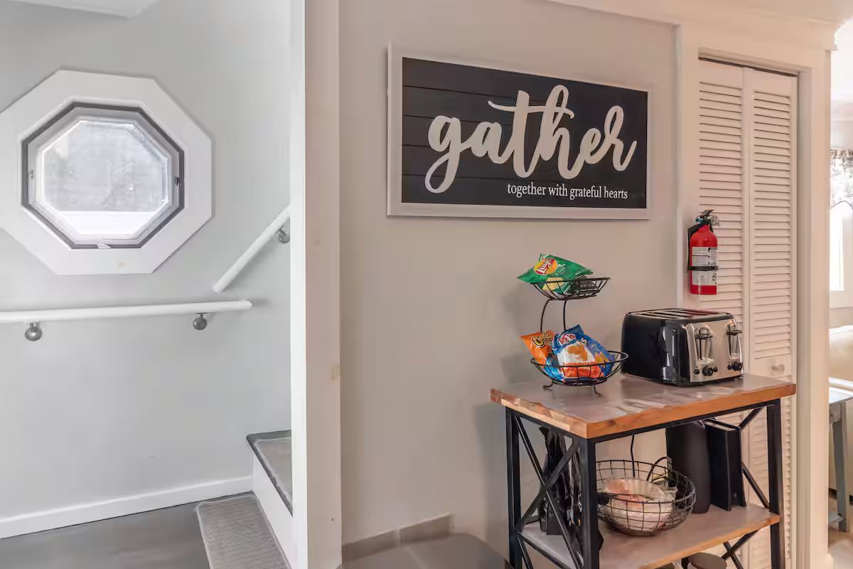 Hallway coffee bar with “gather” sign, toaster, and snacks beside a white stair railing