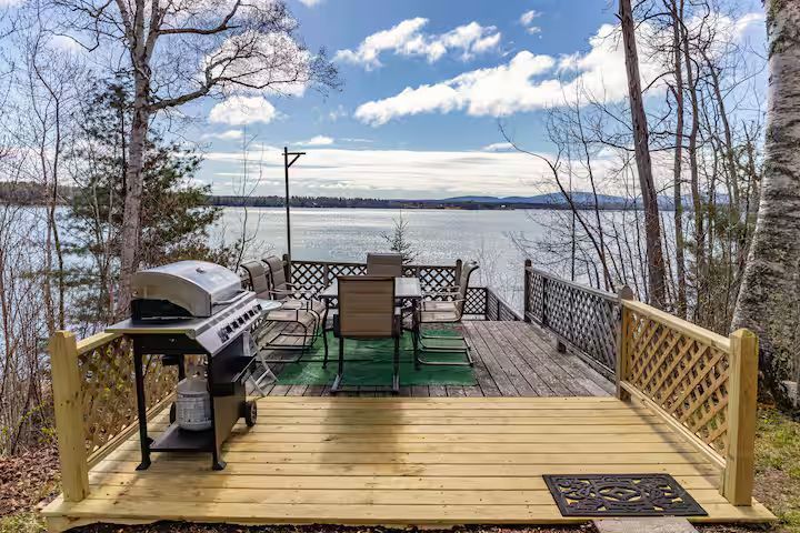 Lakefront deck with grill, dining table, and green outdoor rug under a cloudy sky