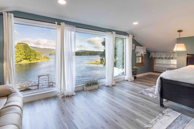 Bright bedroom with floor-to-ceiling windows overlooking a lake and mountains at sunset.