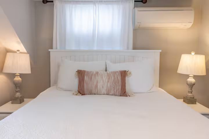 Bright bedroom with a white bed, pink accent pillow, bedside lamps, and a window with curtains