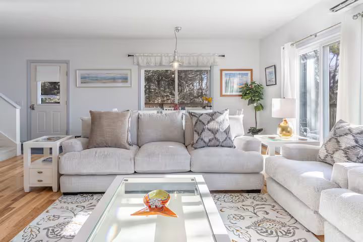 Bright living room with white sofas, patterned rug, coffee table, and large windows